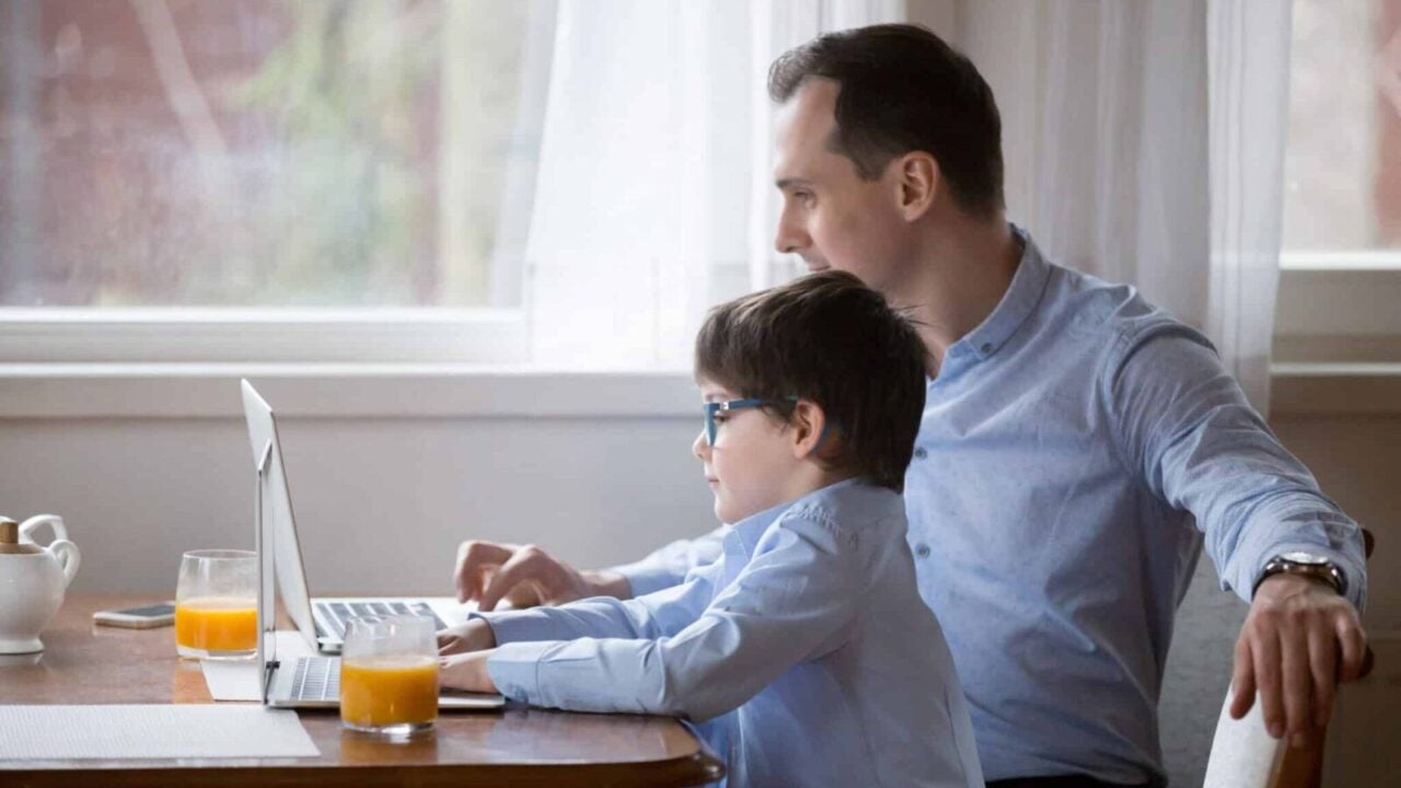 Cute little boy copy dad behavior working at laptop, parent and child sit together in kitchen using computers, father and son have fun enjoy time, small funny kid act like daddy busy with gadget