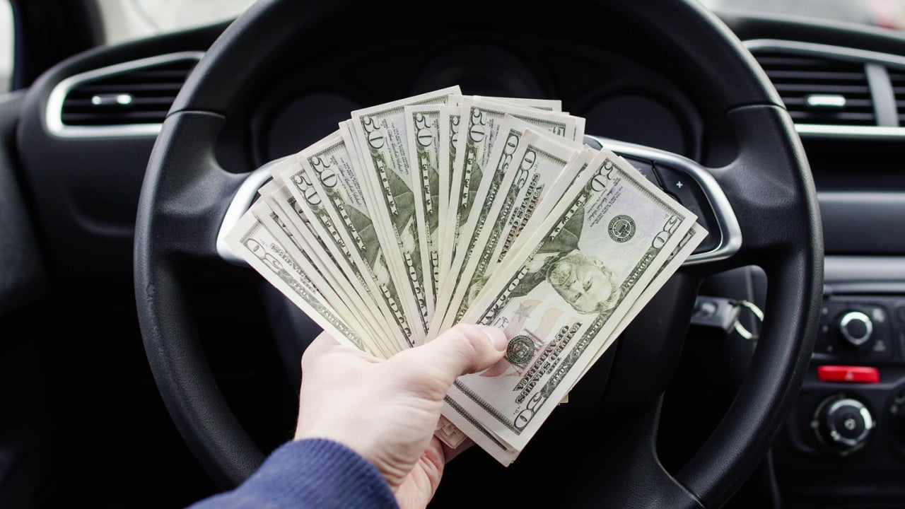 A dollar bills in the male hand on the background of the steering wheel in the car. Concept of earnings or bribes, insurance or credit, investing or selling a car