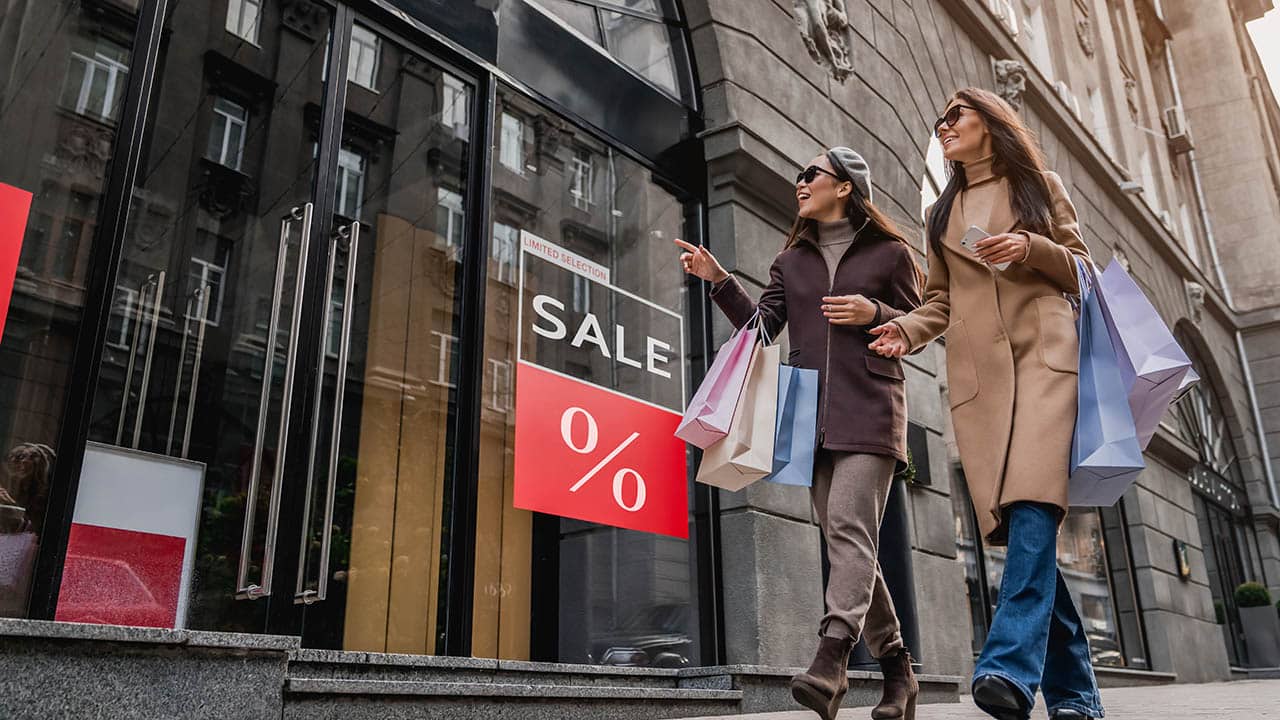 Friends female women walking with shopping bag near Sale sign on shop showcase. Total discount sale, low prices on black friday