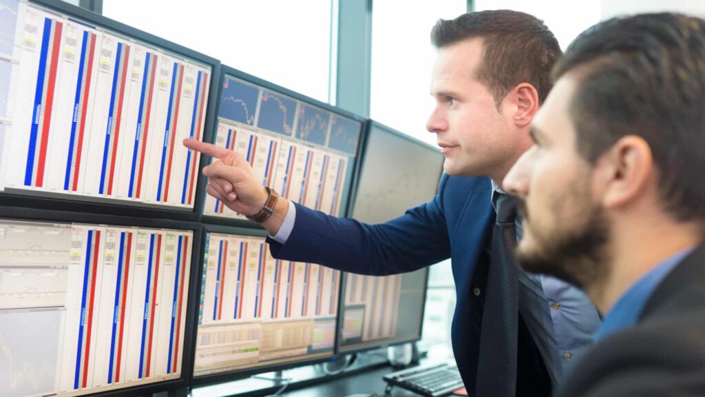 Businessmen trading stocks. Stock traders looking at graphs, indexes and numbers on multiple computer screens. Colleagues in discussion in traders office.